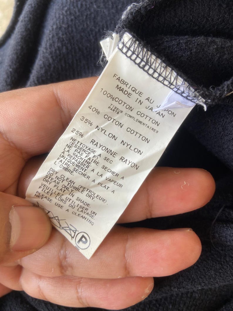 Pu&ograve; includere: Un'etichetta di abbigliamento blu navy con testo in inglese e francese, che descrive la composizione del tessuto e le istruzioni per la cura. L'etichetta recita "FABRIQUE AU JAPON MADE IN JAPAN" ed elenca le percentuali di cotone, nylon e rayon.