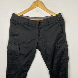 May include: Black cargo trousers with multiple pockets, hanging on a wooden hanger. The trousers have a button closure and a classic design. The fabric appears to be a durable material, suitable for various activities.