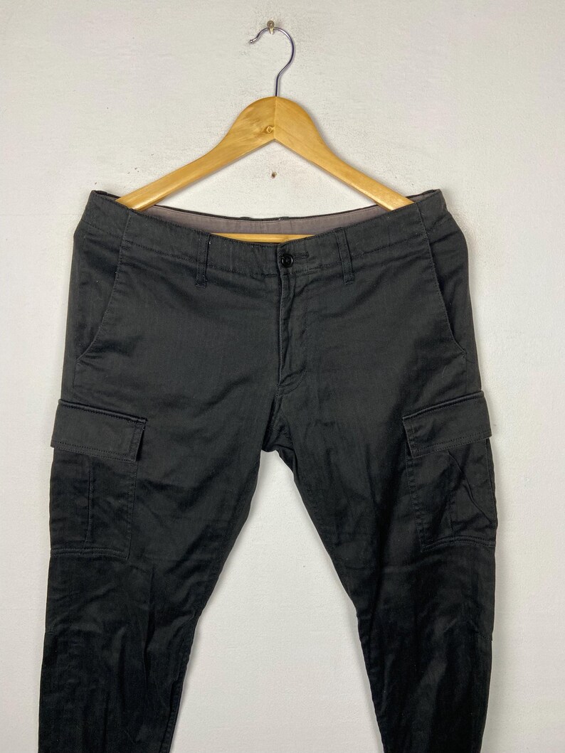 May include: Black cargo trousers with multiple pockets, hanging on a wooden hanger. The trousers have a button closure and a classic design. The fabric appears to be a durable material, suitable for various activities.