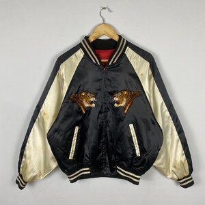 Puede incluir: Chaqueta bomber de sat&eacute;n negro con mangas color crema y cabezas de tigre bordadas. La chaqueta tiene cuello, pu&ntilde;os y dobladillo de canal&eacute; con rayas blancas. Prenda de estilo vintage.
