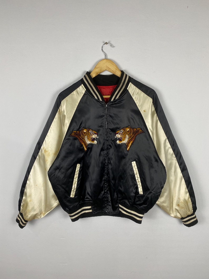 Puede incluir: Chaqueta bomber de sat&eacute;n negro con mangas color crema y cabezas de tigre bordadas. La chaqueta tiene cuello, pu&ntilde;os y dobladillo de canal&eacute; con rayas blancas. Prenda de estilo vintage.