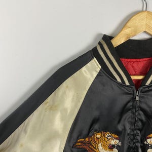 Puede incluir: Chaqueta bomber de sat&eacute;n negro con mangas color crema y forro rojo. La chaqueta presenta un dise&ntilde;o de tigre bordado en tonos marr&oacute;n, naranja y blanco. El cuello es negro con rayas blancas. Una cremallera recorre la parte delantera.