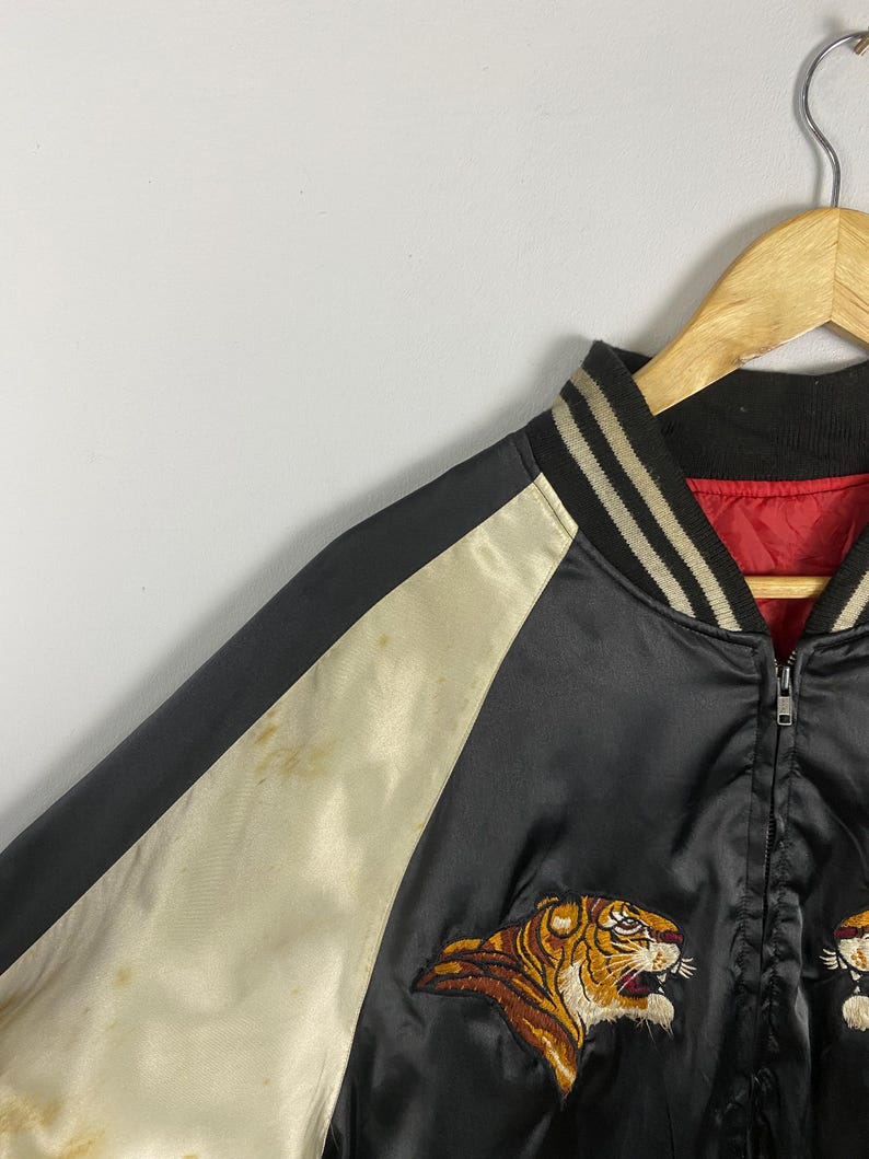 Puede incluir: Chaqueta bomber de sat&eacute;n negro con mangas color crema y forro rojo. La chaqueta presenta un dise&ntilde;o de tigre bordado en tonos marr&oacute;n, naranja y blanco. El cuello es negro con rayas blancas. Una cremallera recorre la parte delantera.