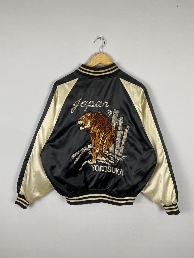 Puede incluir: Chaqueta bomber de sat&eacute;n negro con mangas color crema. La espalda presenta un tigre bordado, bamb&uacute; y las palabras "Japan" y "Yokosuka". La chaqueta tiene cuello, pu&ntilde;os y dobladillo acanalados.