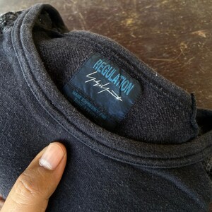 Pu&ograve; includere: Maglietta girocollo grigio scuro con un'etichetta che recita "REGULATION Yohji Yamamoto" in blu e bianco. L'etichetta include anche il testo "MADE EXPRESSLY FOR YOHJI'S PEOPLE". La maglietta &egrave; realizzata in un tessuto morbido e strutturato.