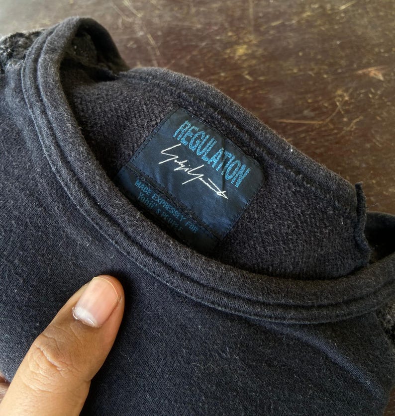 Pu&ograve; includere: Maglietta girocollo grigio scuro con un'etichetta che recita "REGULATION Yohji Yamamoto" in blu e bianco. L'etichetta include anche il testo "MADE EXPRESSLY FOR YOHJI'S PEOPLE". La maglietta &egrave; realizzata in un tessuto morbido e strutturato.