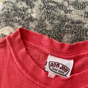 May include: Close-up of a coral-colored t-shirt with a crew neck. The shirt features a tag with the text "RON JON SURF SHOP" in a red and white oval design. The fabric appears slightly worn.