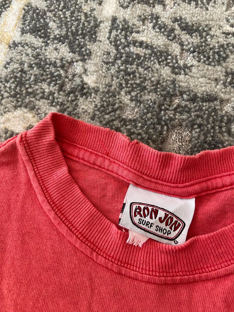 May include: Close-up of a coral-colored t-shirt with a crew neck. The shirt features a tag with the text "RON JON SURF SHOP" in a red and white oval design. The fabric appears slightly worn.
