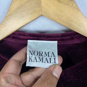 以下が含まれることがあります： 「Norma Kamali」と書いてある白いタグが付いた、濃い赤紫色のベルベットのシャツのクローズアップ。