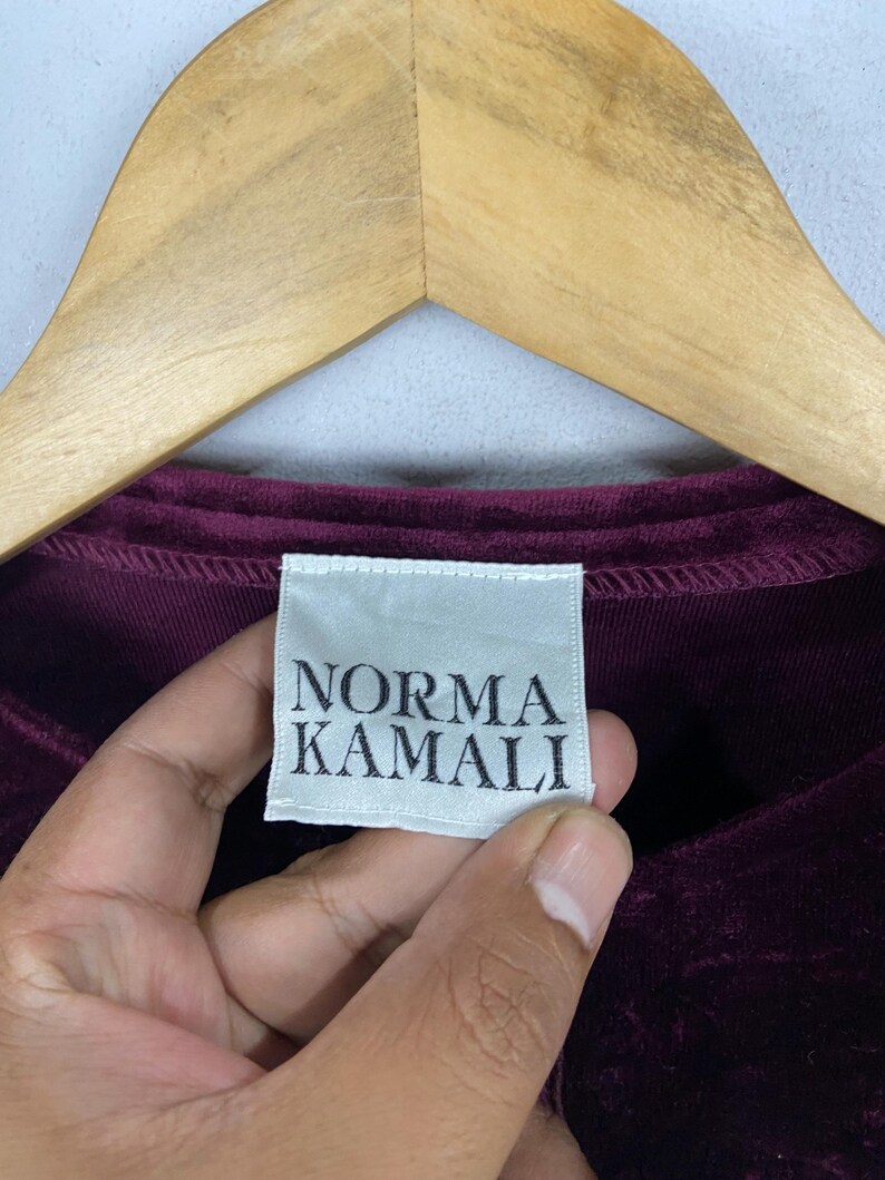 以下が含まれることがあります： 「Norma Kamali」と書いてある白いタグが付いた、濃い赤紫色のベルベットのシャツのクローズアップ。