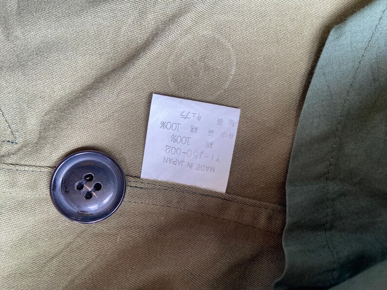 May include: Close-up of olive green fabric with a dark button and a white tag. The tag has Japanese text and reads "MADE IN JAPAN". The button is round with four holes. The fabric appears to be cotton or a similar material.