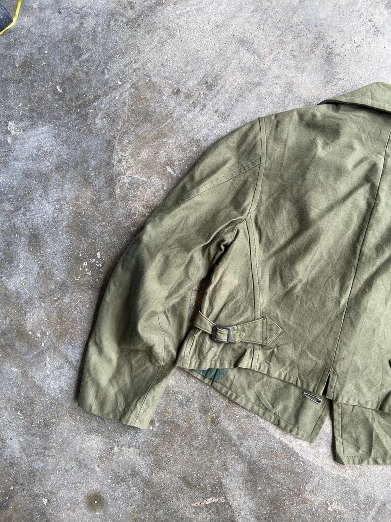 May include: Olive green jacket with a collar and a belt detail at the waist. The jacket has a classic design with a tailored fit. The fabric appears to be a durable material, suitable for various weather conditions.