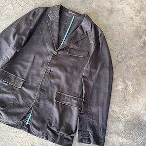 May include: A dark brown blazer featuring a notched lapel, three front buttons, and two large flap pockets at the waist. The garment includes a single chest pocket and a small shoulder epaulet detail, with a bright turquoise lining visible along the inner placket.