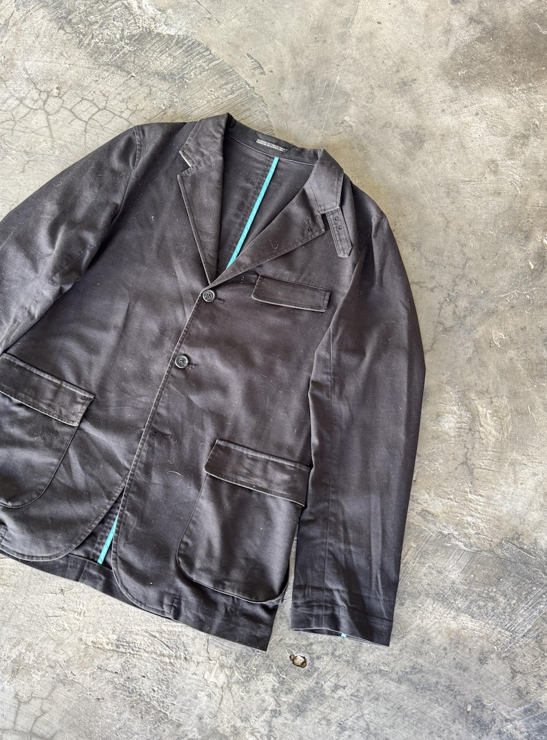 May include: A dark brown blazer featuring a notched lapel, three front buttons, and two large flap pockets at the waist. The garment includes a single chest pocket and a small shoulder epaulet detail, with a bright turquoise lining visible along the inner placket.