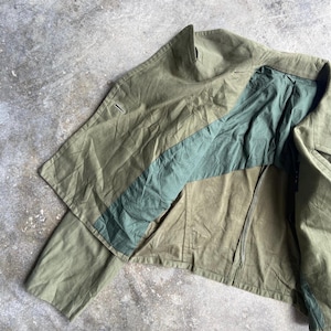 May include: Olive green jacket with a contrasting teal stripe across the chest. The jacket has a zipper closure and a high collar. The fabric appears to be a durable, possibly canvas material. The jacket is laid out on a concrete surface.