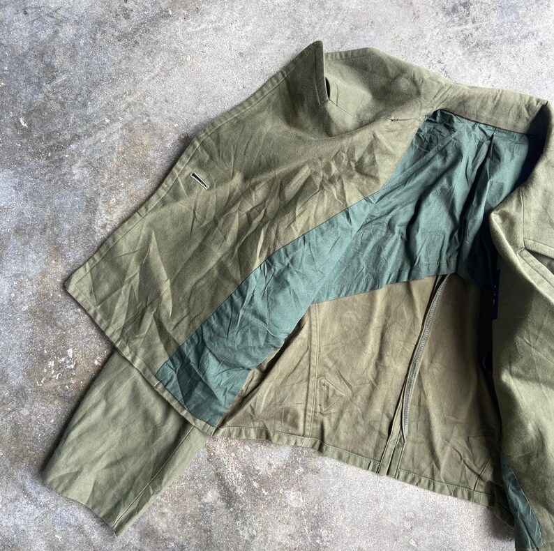 May include: Olive green jacket with a contrasting teal stripe across the chest. The jacket has a zipper closure and a high collar. The fabric appears to be a durable, possibly canvas material. The jacket is laid out on a concrete surface.