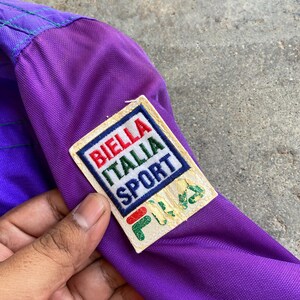 May include: Close-up of a vintage purple jacket with a Biella Italia Sport patch. The patch features red, white, and green text and a green logo. The jacket has a purple and red colour scheme.