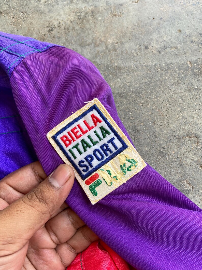 May include: Close-up of a vintage purple jacket with a Biella Italia Sport patch. The patch features red, white, and green text and a green logo. The jacket has a purple and red colour scheme.