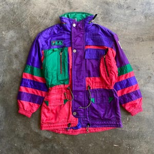 May include: A vintage Fila ski jacket in a vibrant colour block design. The jacket features purple, green, and pink panels with the Fila logo. It has multiple pockets, a drawstring waist, and a high collar.