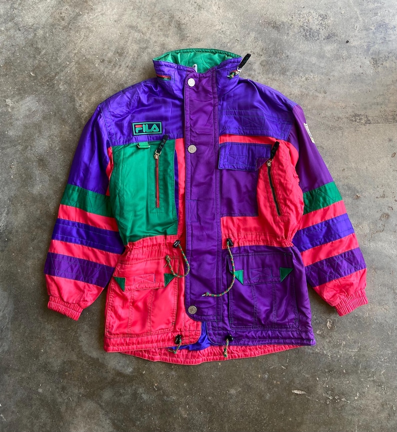 May include: A vintage Fila ski jacket in a vibrant colour block design. The jacket features purple, green, and pink panels with the Fila logo. It has multiple pockets, a drawstring waist, and a high collar.