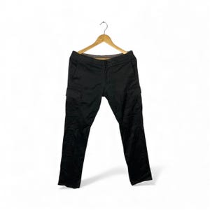 May include: Black cargo trousers hanging on a wooden hanger. The trousers feature two side pockets and a straight leg cut. The material looks durable, suitable for outdoor activities or everyday wear. The trousers are approximately 100 cm in length.