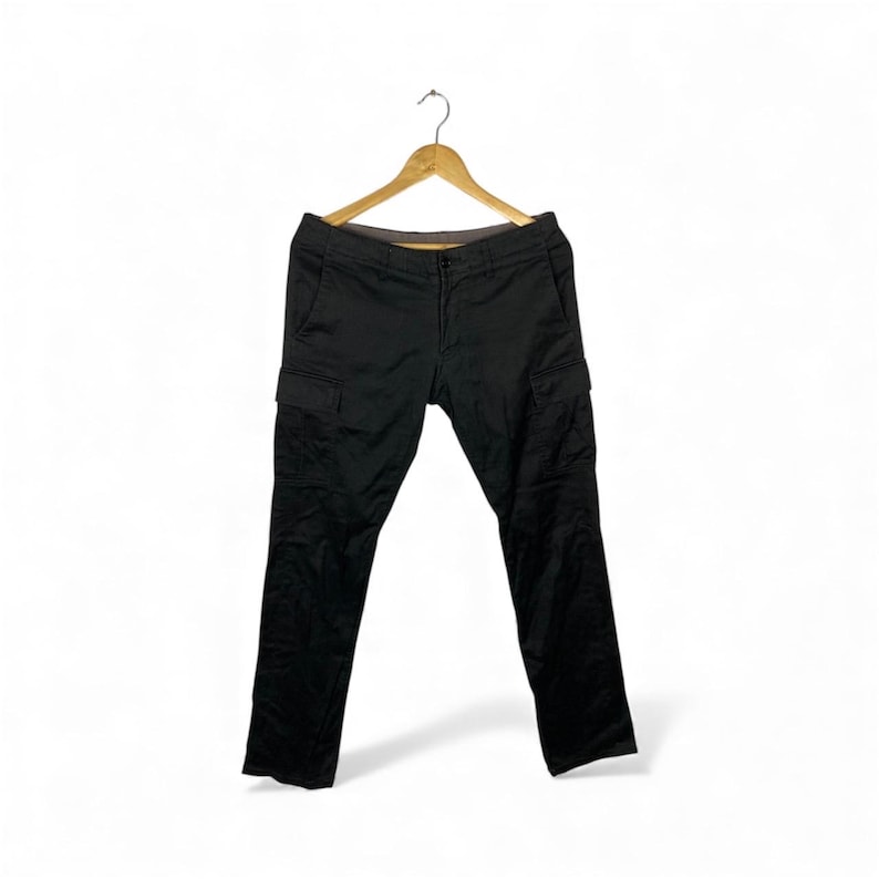 May include: Black cargo trousers hanging on a wooden hanger. The trousers feature two side pockets and a straight leg cut. The material looks durable, suitable for outdoor activities or everyday wear. The trousers are approximately 100 cm in length.