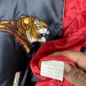 Puede incluir: Chaqueta de sat&eacute;n azul marino con un dise&ntilde;o de tigre bordado en tonos dorados, marrones y blancos. La chaqueta tiene un forro rojo y un ribete blanco. Se ve una etiqueta con texto japon&eacute;s.