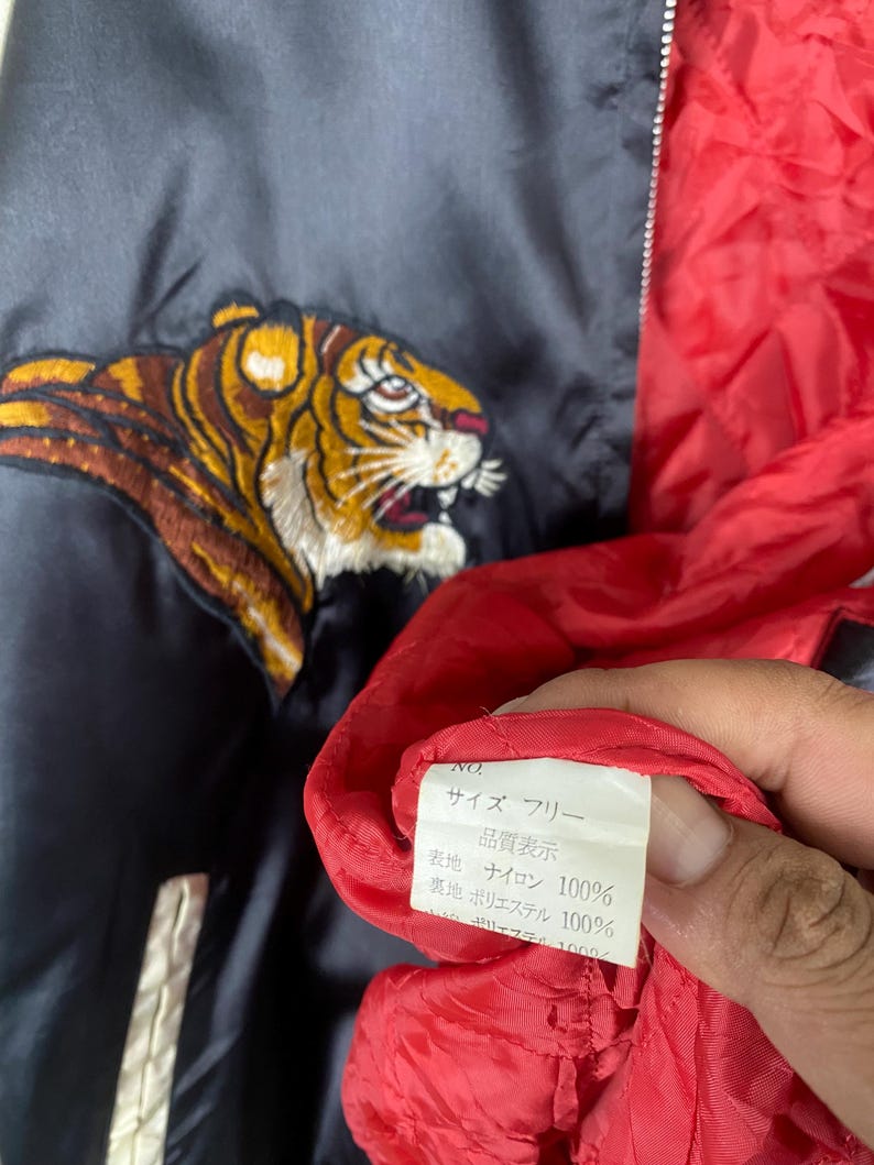 Puede incluir: Chaqueta de sat&eacute;n azul marino con un dise&ntilde;o de tigre bordado en tonos dorados, marrones y blancos. La chaqueta tiene un forro rojo y un ribete blanco. Se ve una etiqueta con texto japon&eacute;s.