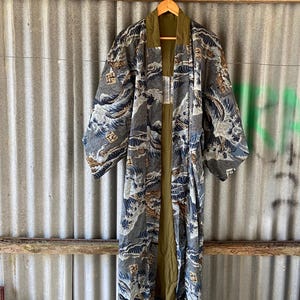 Puede incluir: Larga bata tipo kimono abierta por delante con un estampado de olas en tonos grises, azules y marrones. La bata tiene mangas anchas y un forro verde. Cuelga de una percha de madera sobre un fondo de metal corrugado.
