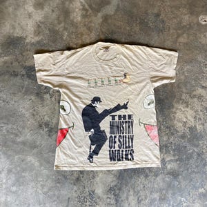 May include: A beige short-sleeve t-shirt featuring a graphic of a person in a suit performing a high kick with the text The Ministry of Silly Walks. Above this, a row of small figures and the text Monty Python's Flying Circus are printed.