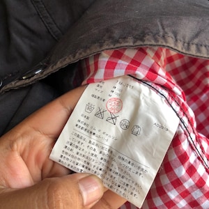 May include: Close-up of a dark brown jacket interior with a red and white checkered lining. A white tag with Japanese text and care symbols is visible. The tag includes the text "1M-J011" and "AD2013".