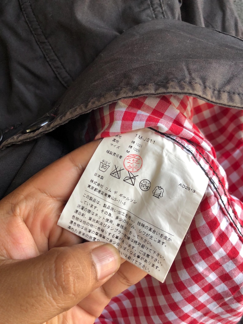 May include: Close-up of a dark brown jacket interior with a red and white checkered lining. A white tag with Japanese text and care symbols is visible. The tag includes the text "1M-J011" and "AD2013".