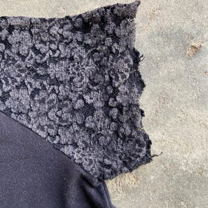 Pu&ograve; includere: Primo piano di un capo nero con una manica in pizzo grigio. Il pizzo presenta un motivo floreale ed &egrave; attaccato a un tessuto scuro. La manica &egrave; angolata, evidenziando il contrasto delle trame.