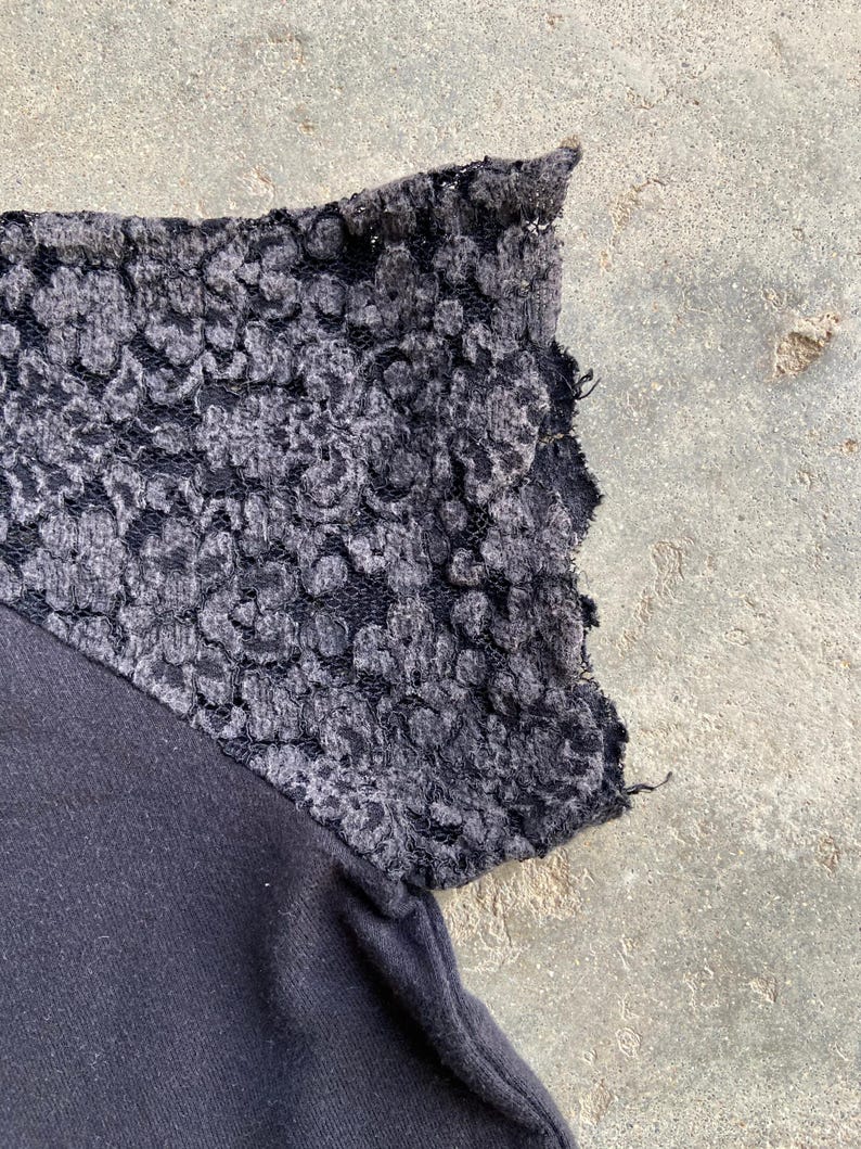 Pu&ograve; includere: Primo piano di un capo nero con una manica in pizzo grigio. Il pizzo presenta un motivo floreale ed &egrave; attaccato a un tessuto scuro. La manica &egrave; angolata, evidenziando il contrasto delle trame.
