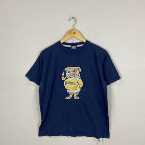 May include: A navy blue t-shirt featuring a graphic of a bulldog wearing a yellow sweater and smoking a pipe. The text "Pharaohs" is printed on the sweater.