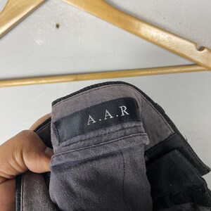 May include: Close-up of grey trousers with a black waistband. The label reads "A.A.R" in white lettering. The trousers are hanging on a wooden hanger.