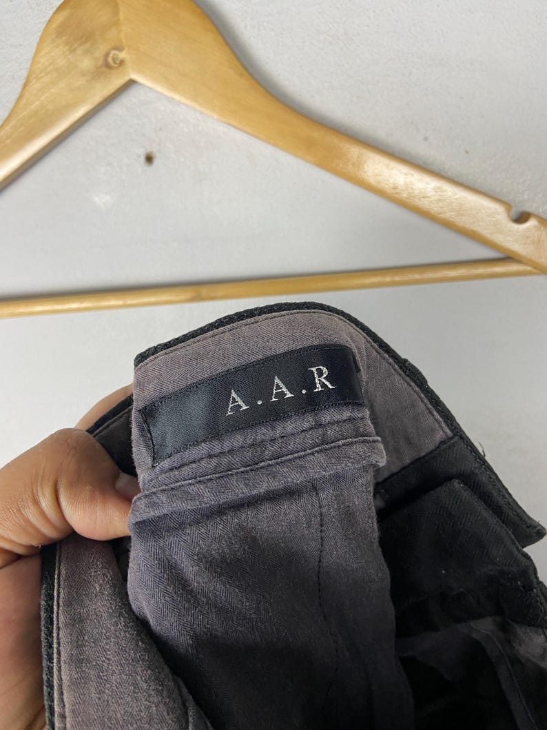 May include: Close-up of grey trousers with a black waistband. The label reads "A.A.R" in white lettering. The trousers are hanging on a wooden hanger.