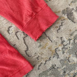 May include: Close-up of two coral-colored long-sleeved shirt cuffs. The cuffs have a ribbed texture and a raw, frayed edge. The fabric appears soft and slightly worn. The background is a patterned rug.