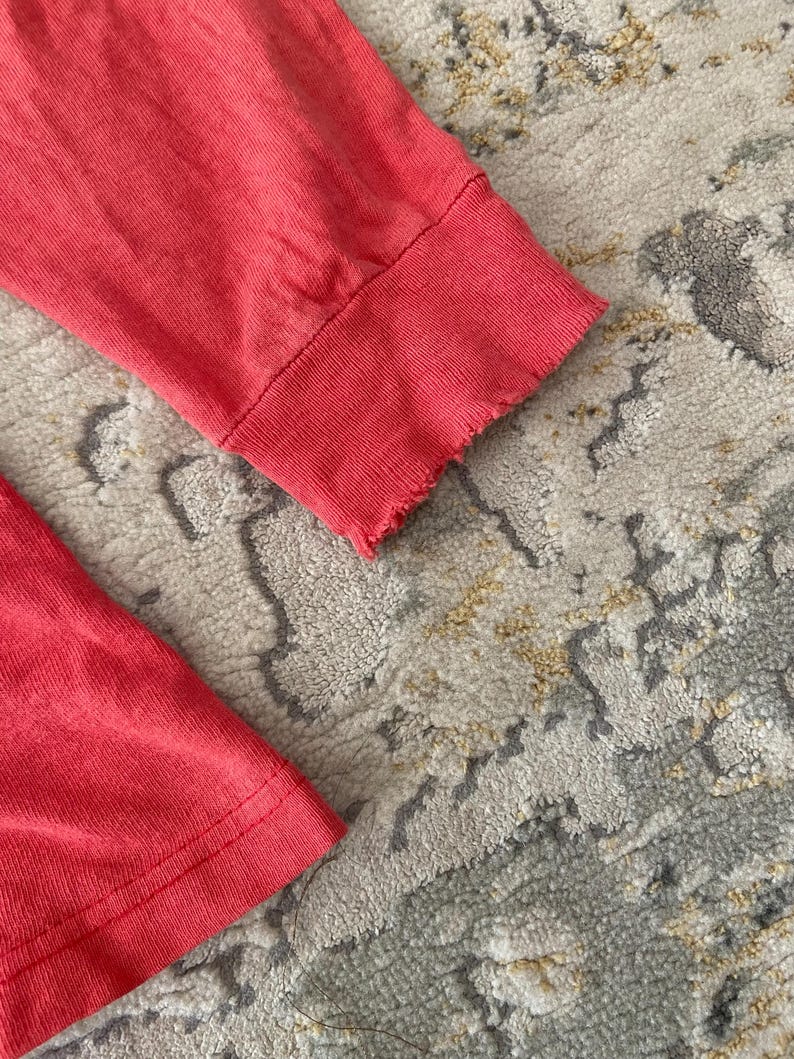 May include: Close-up of two coral-colored long-sleeved shirt cuffs. The cuffs have a ribbed texture and a raw, frayed edge. The fabric appears soft and slightly worn. The background is a patterned rug.