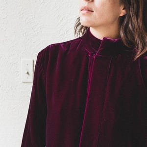 以下が含まれることがあります： ハイネックで首元にボタン留めのある、長袖の濃いパープルのベルベットシャツです。