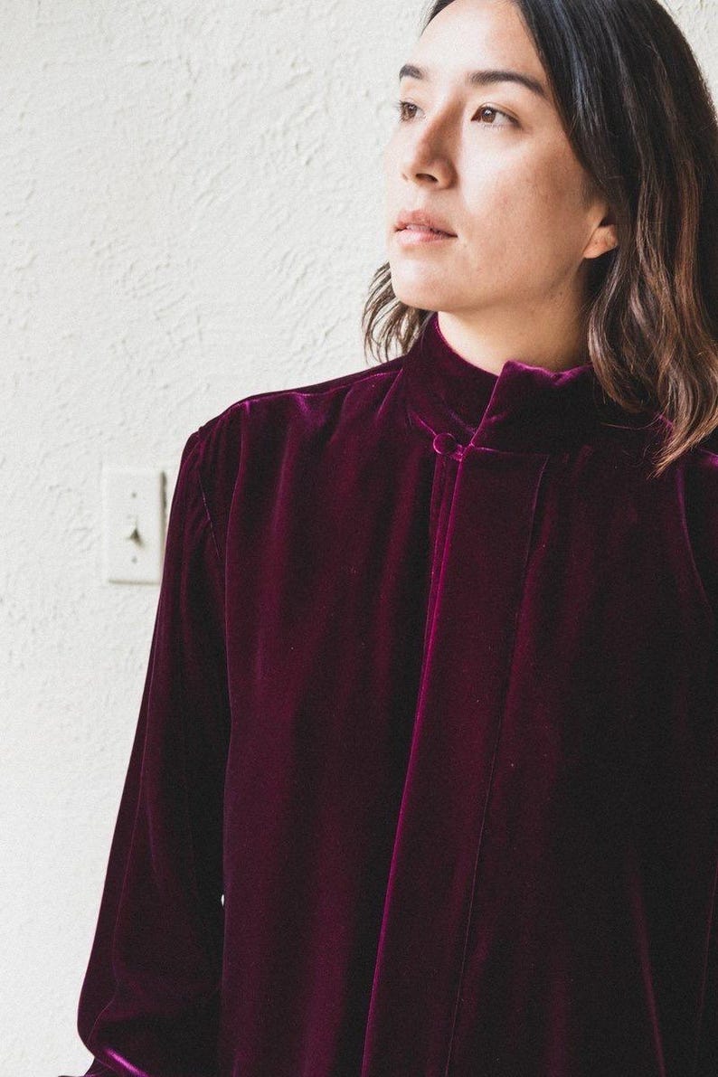 以下が含まれることがあります： ハイネックで首元にボタン留めのある、長袖の濃いパープルのベルベットシャツです。