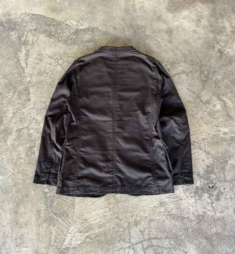 May include: A solid black blazer featuring a structured collar, long sleeves, and a central vertical seam running down the back. The garment is constructed with visible stitching along the shoulders and side panels, showcasing a clean, tailored design.