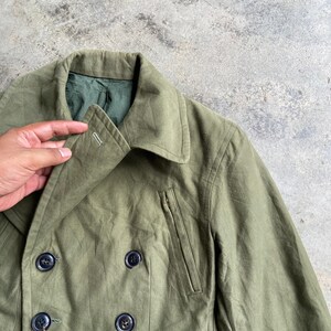 May include: Olive green double-breasted jacket with black buttons. The jacket features a notched collar, a small pocket with a zipper, and a flap pocket. The jacket is made of a durable fabric and has a classic design.