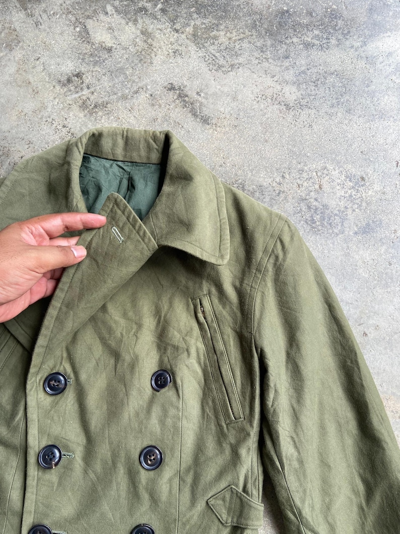 May include: Olive green double-breasted jacket with black buttons. The jacket features a notched collar, a small pocket with a zipper, and a flap pocket. The jacket is made of a durable fabric and has a classic design.