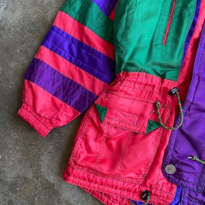 May include: A vintage colour block jacket in shades of purple, green, and red. The jacket features a zip-up front, pockets, and a drawstring. The sleeves have a striped pattern. The jacket is made of a lightweight material.