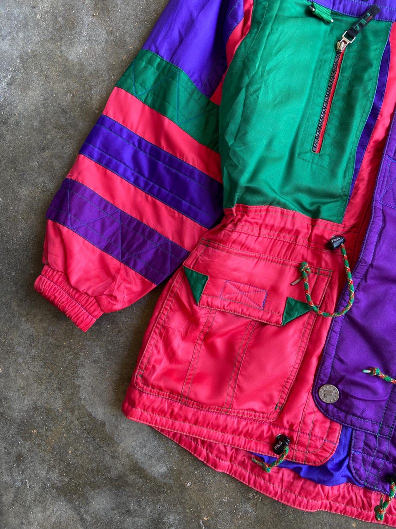 May include: A vintage colour block jacket in shades of purple, green, and red. The jacket features a zip-up front, pockets, and a drawstring. The sleeves have a striped pattern. The jacket is made of a lightweight material.