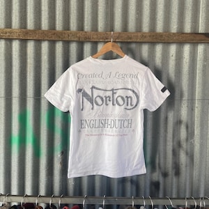 May include: White t-shirt featuring the text "Norton" in a distinctive font, with additional text including "Created A Legend" and "ENGLISH-DUTCH". The shirt hangs on a wooden hanger against a corrugated metal backdrop.