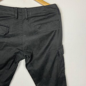 May include: Black cargo trousers with multiple pockets, including two back pockets and two side cargo pockets. The trousers are hanging on a wooden hanger against a white background. The trousers are made of a durable fabric.