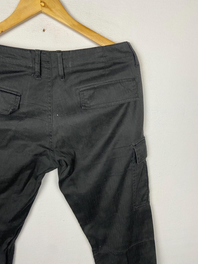 May include: Black cargo trousers with multiple pockets, including two back pockets and two side cargo pockets. The trousers are hanging on a wooden hanger against a white background. The trousers are made of a durable fabric.