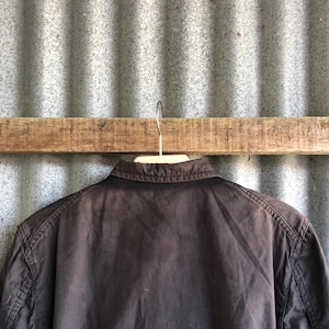 May include: A dark brown jacket hanging on a wooden bar. The jacket has a collar and visible stitching along the seams. The background is a corrugated metal sheet.